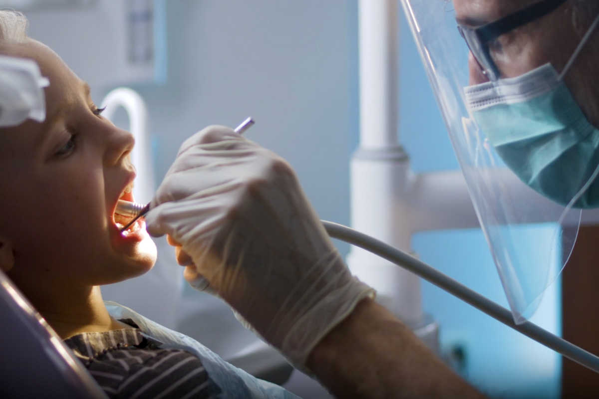 Dentist wearing gloves, mask, and face shield performing a dental examination on a child in a clinic