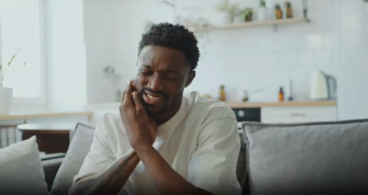 Person on a couch clutching their cheek, wincing while seeking tooth pain relief at home.