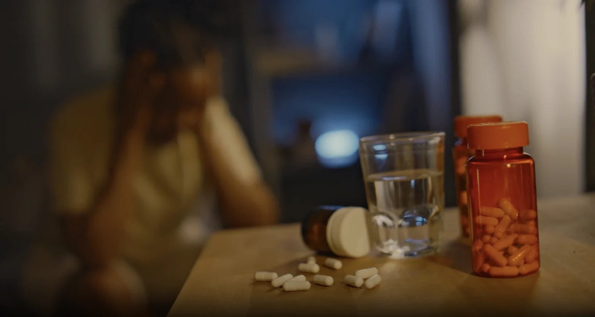 Medication bottles, spilled pills, and a glass of water beside a distressed person at a table.