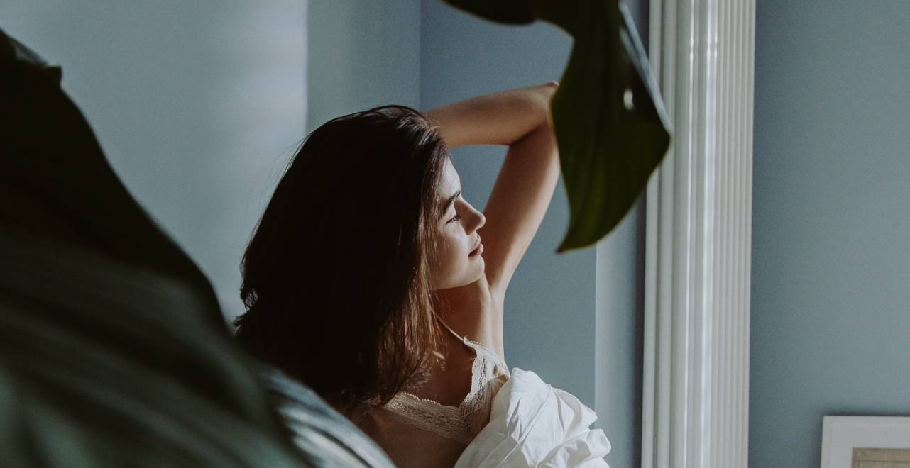 Woman basking in soft natural light near a window, surrounded by leafy plants & serene ambiance