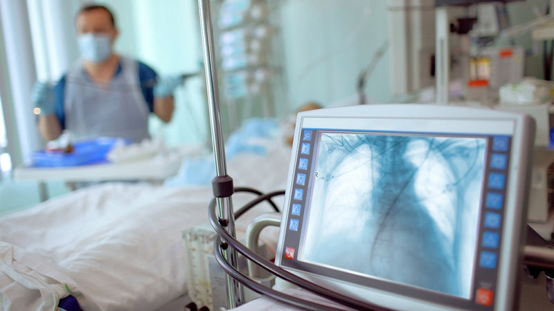 Chest X-ray on a hospital monitor as a masked clinician tends a patient in the ICU.