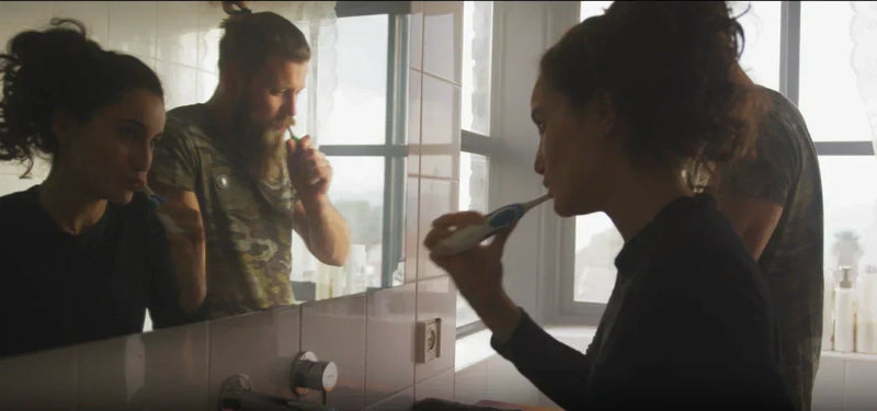 Couple brushing their teeth together at a bathroom mirror in soft morning light.