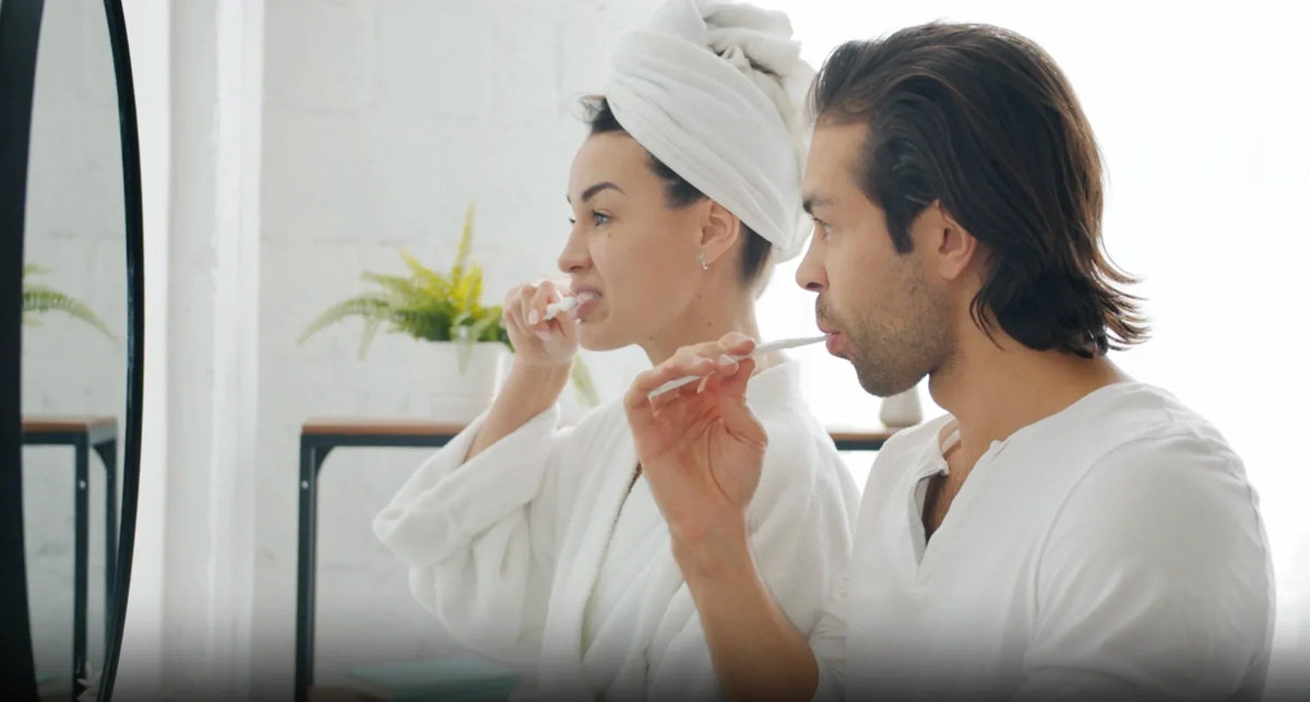 Two people in white robes brushing their teeth together at a bright bathroom mirror.