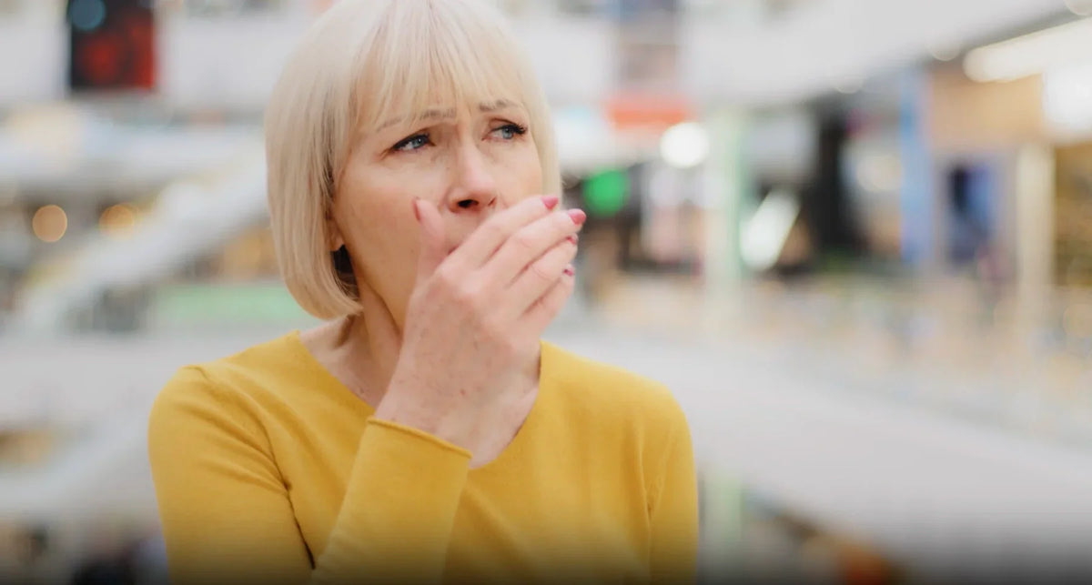 Person in yellow top covering mouth with hand, worried about bad breath in a mall.