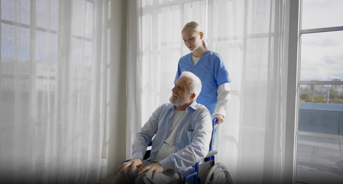 Nurse in blue scrubs assisting a senior man in a wheelchair beside sunlit windows in senior care.