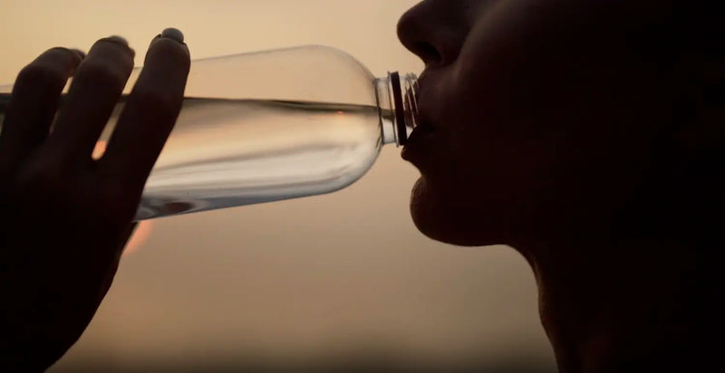 Person drinking water from a clear bottle at sunset, illustrating hydration for oral health.