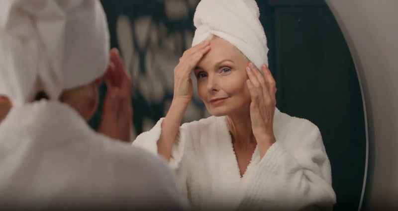 Person in white robe and hair towel checking facial skin for signs of aging in mirror.