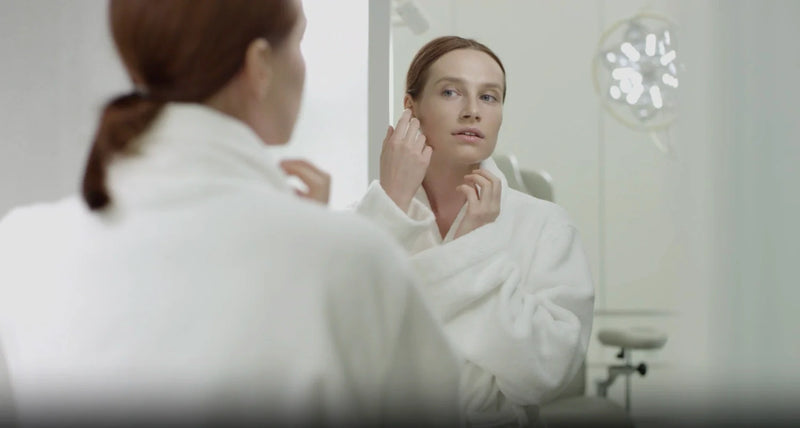 Person in a white robe examines facial skin in a mirror, reflecting collagen supplements for skin.