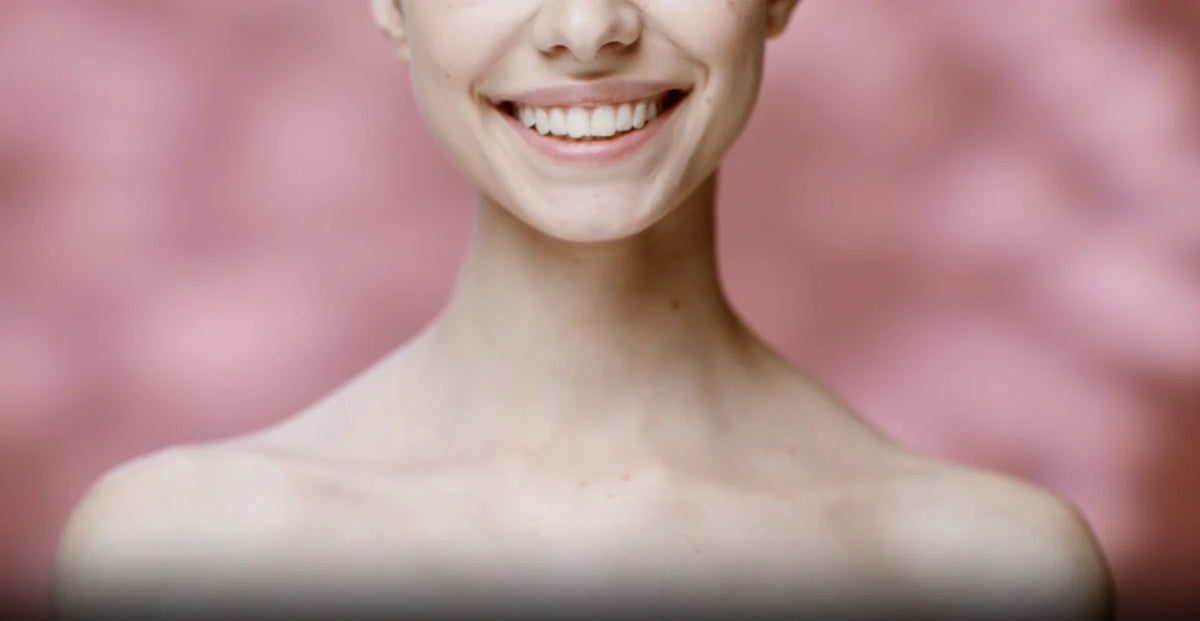 Close-up smile with clear skin and bare shoulders against a soft pink backdrop.