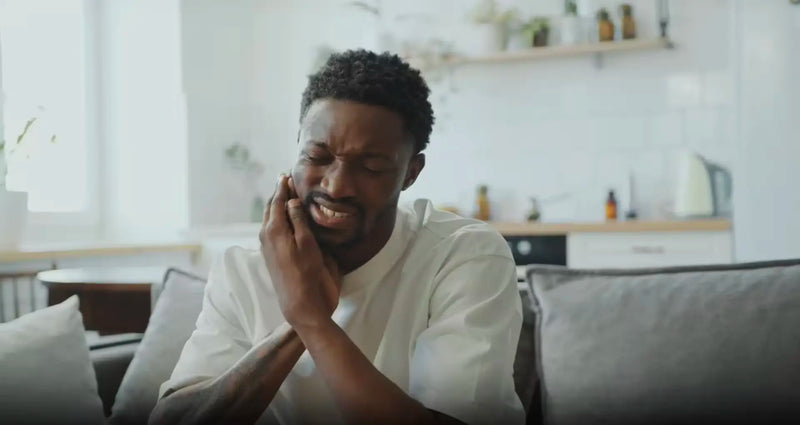 Person on a couch clutching their cheek, wincing while seeking tooth pain relief at home.