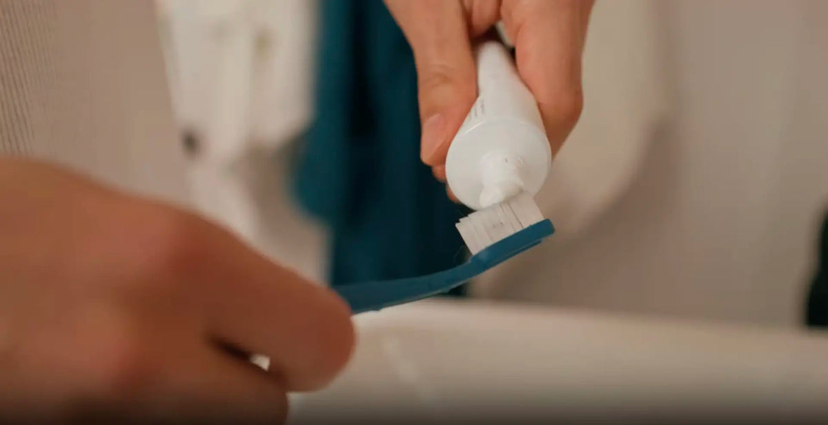 Hands squeezing toothpaste onto a blue manual toothbrush with soft bristles.