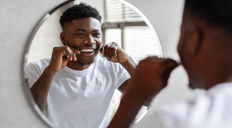 Person flossing teeth while smiling into a round bathroom mirror by a window.