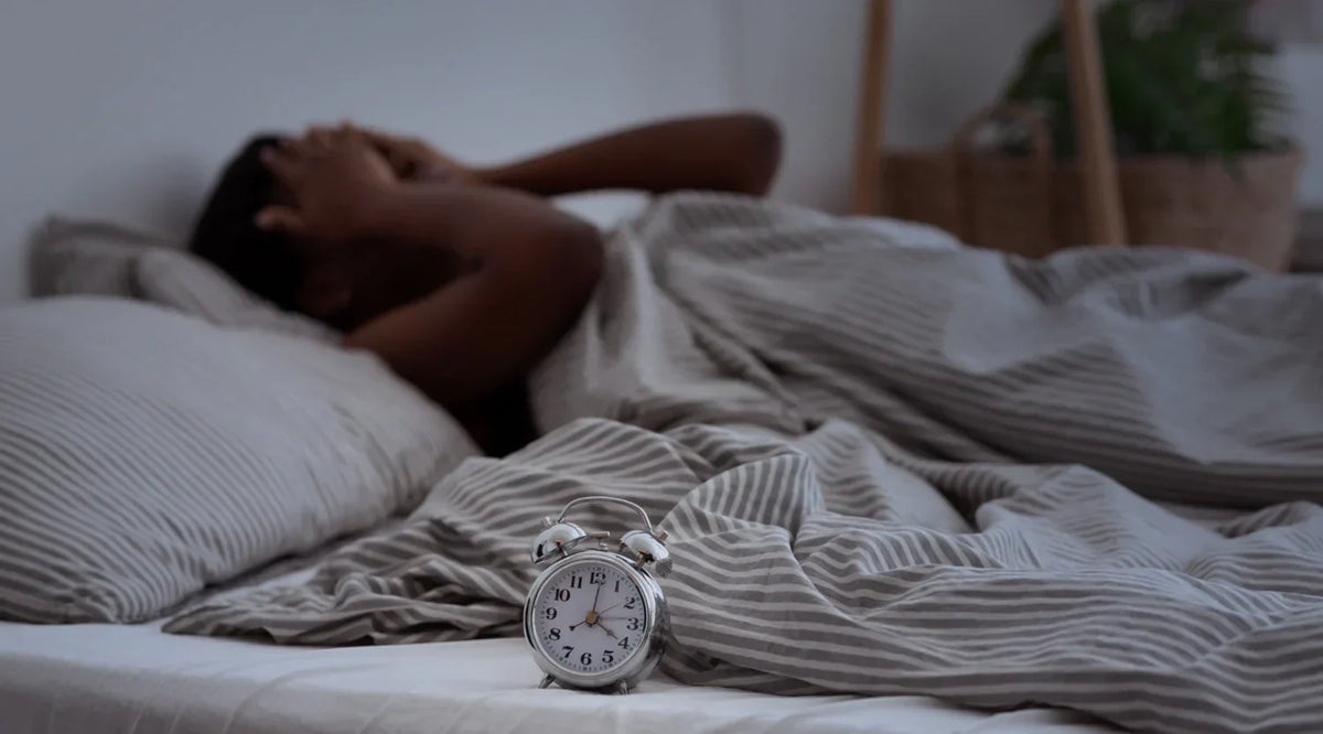 Person lying awake in bed beside an alarm clock, struggling with restless sleep.