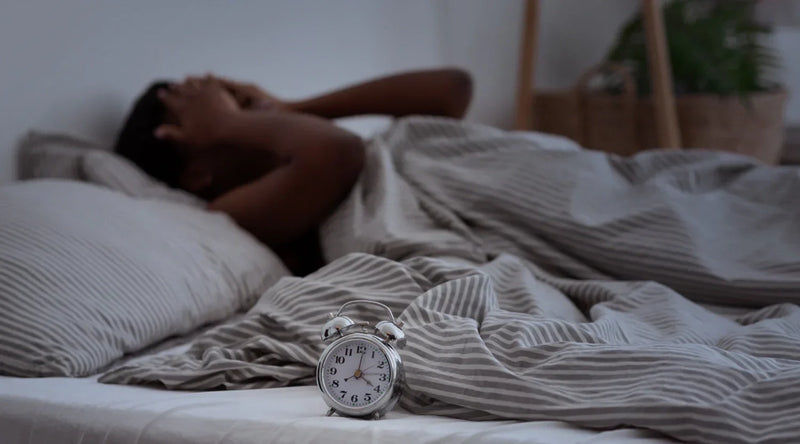 Person lying awake in bed beside an alarm clock, struggling with restless sleep.