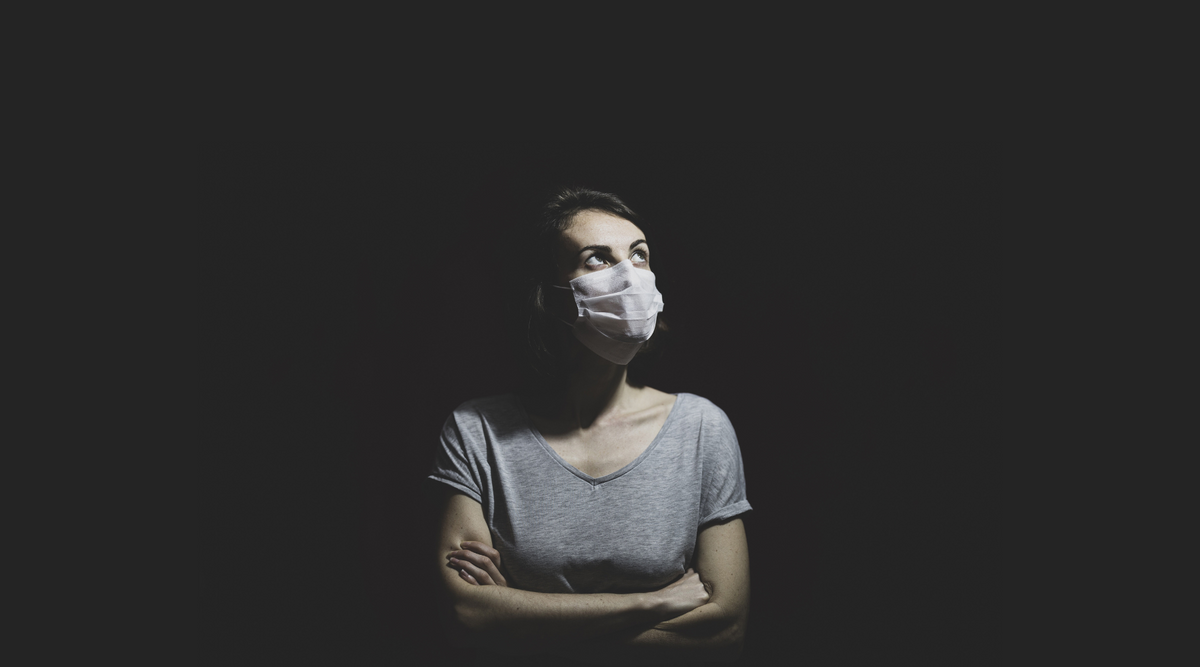 Person in a surgical mask, arms crossed, looking upward against a dark background.