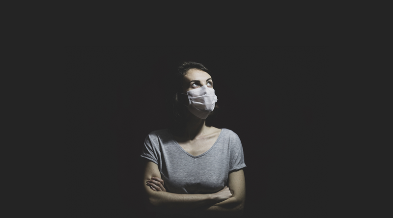 Person in a surgical mask, arms crossed, looking upward against a dark background.