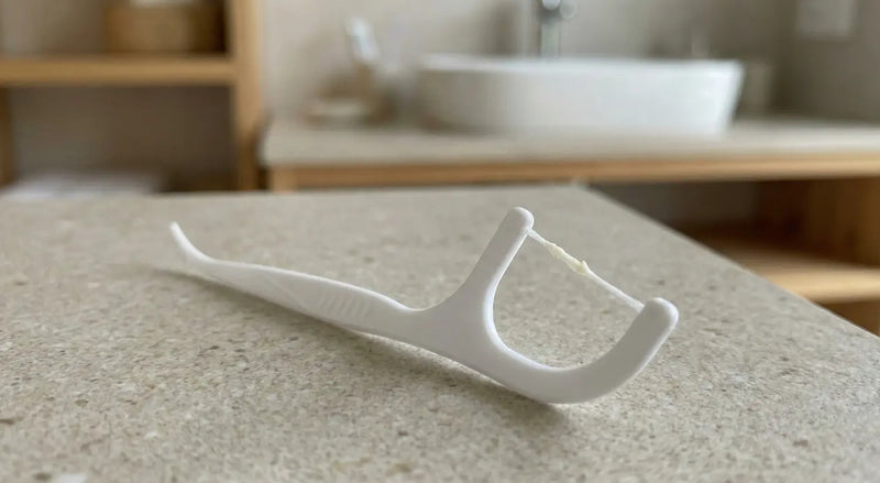 White plastic floss pick with used floss resting on a bathroom countertop near a sink.