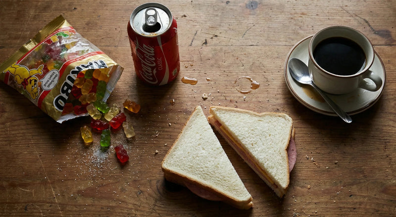 Spilled gummy bears, cola can, white bread sandwich, and black coffee on a wooden table.