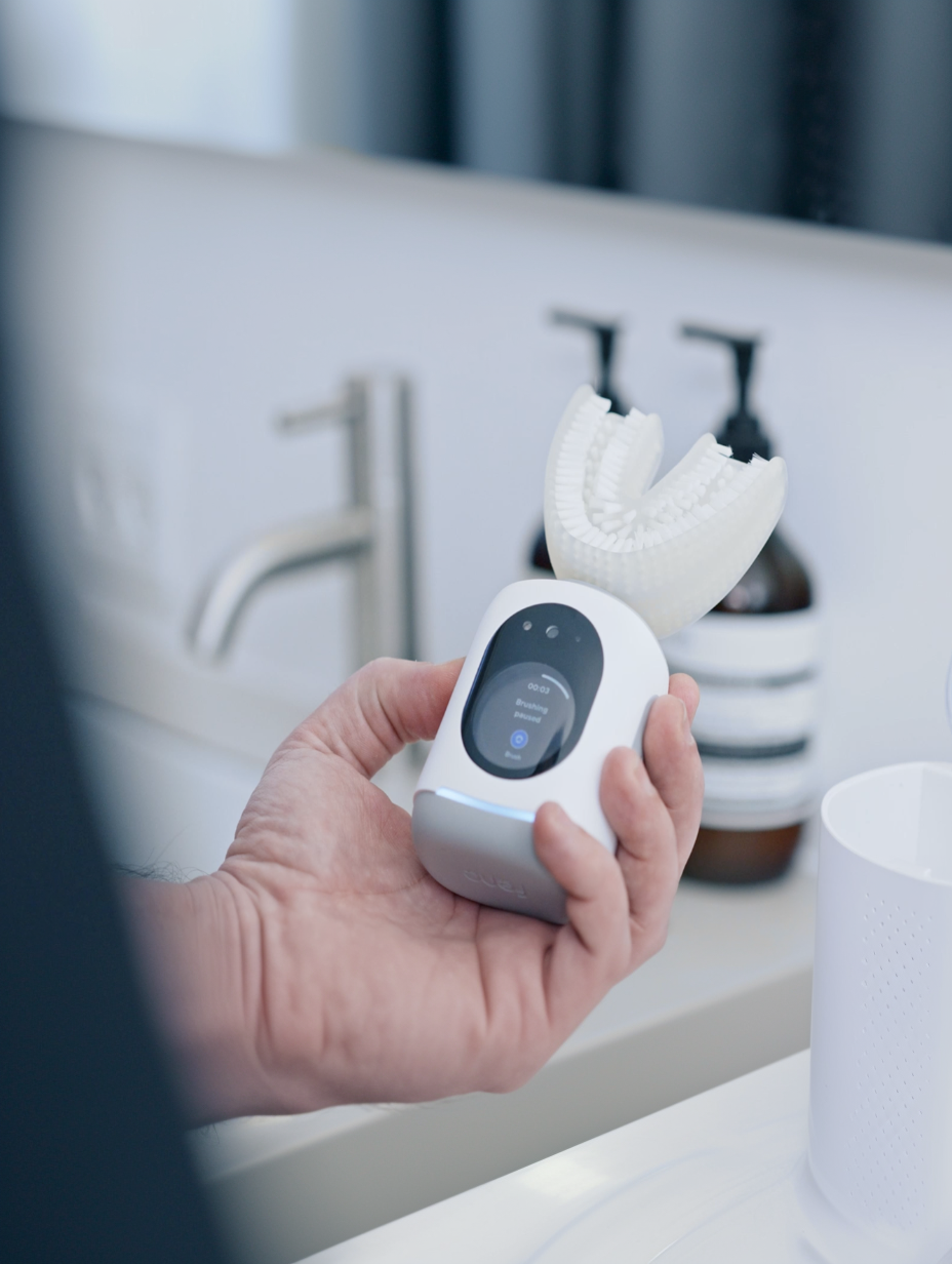 Hand holding Feno U-shaped smart toothbrush with lit screen at a bathroom sink.