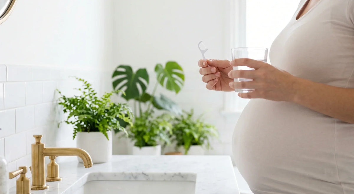 Pregnant person holding a floss pick and glass of water at a bright bathroom sink.