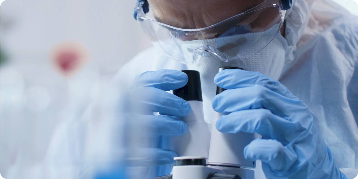 Researcher in protective suit and blue gloves peers into a microscope in a lab.